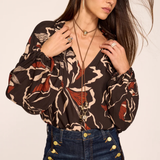 A woman wearing a brown long-sleeve blouse with a beige, black, and rust-red abstract pattern, paired with dark blue jeans featuring gold button details on the pocket, set against a plain beige background.