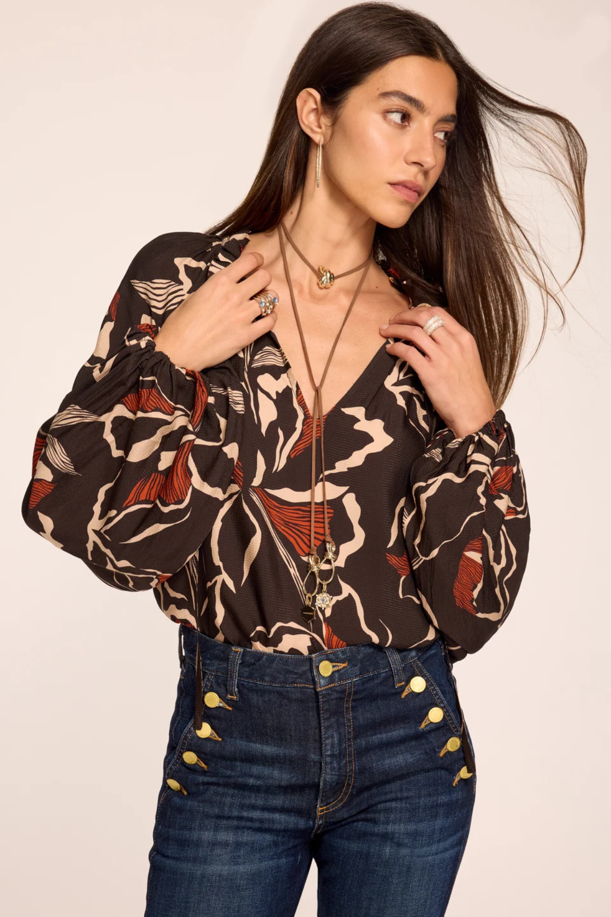 A woman wearing a brown long-sleeve blouse with a beige, black, and rust-red abstract pattern, paired with dark blue jeans featuring gold button details on the pocket, set against a plain beige background.