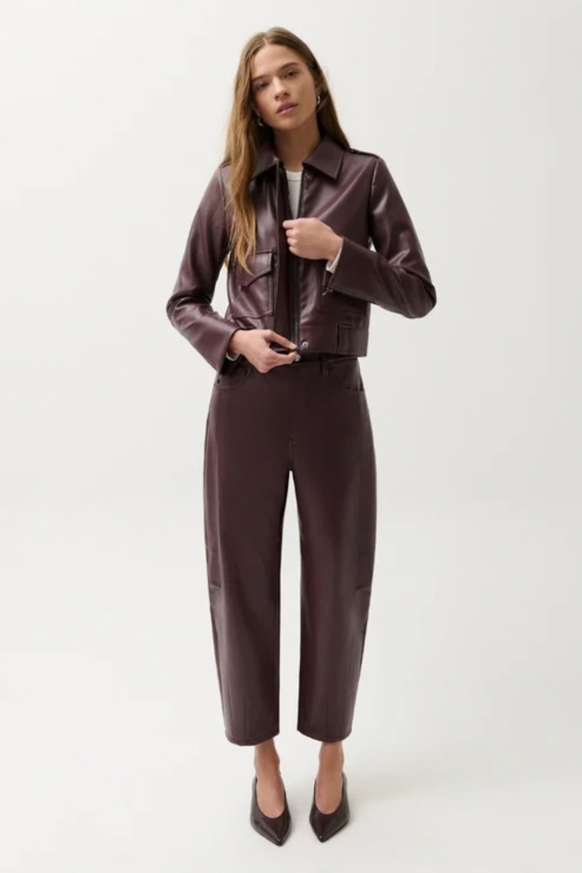 A woman modeling a dark brown faux-leather jacket and matching wide-leg cropped trousers against a plain light gray studio background.