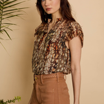 A woman modeling a short-sleeve button-up blouse with an abstract animal-like print in brown, beige, and orange tones, paired with brown pants, set against a warm beige background with green plants.