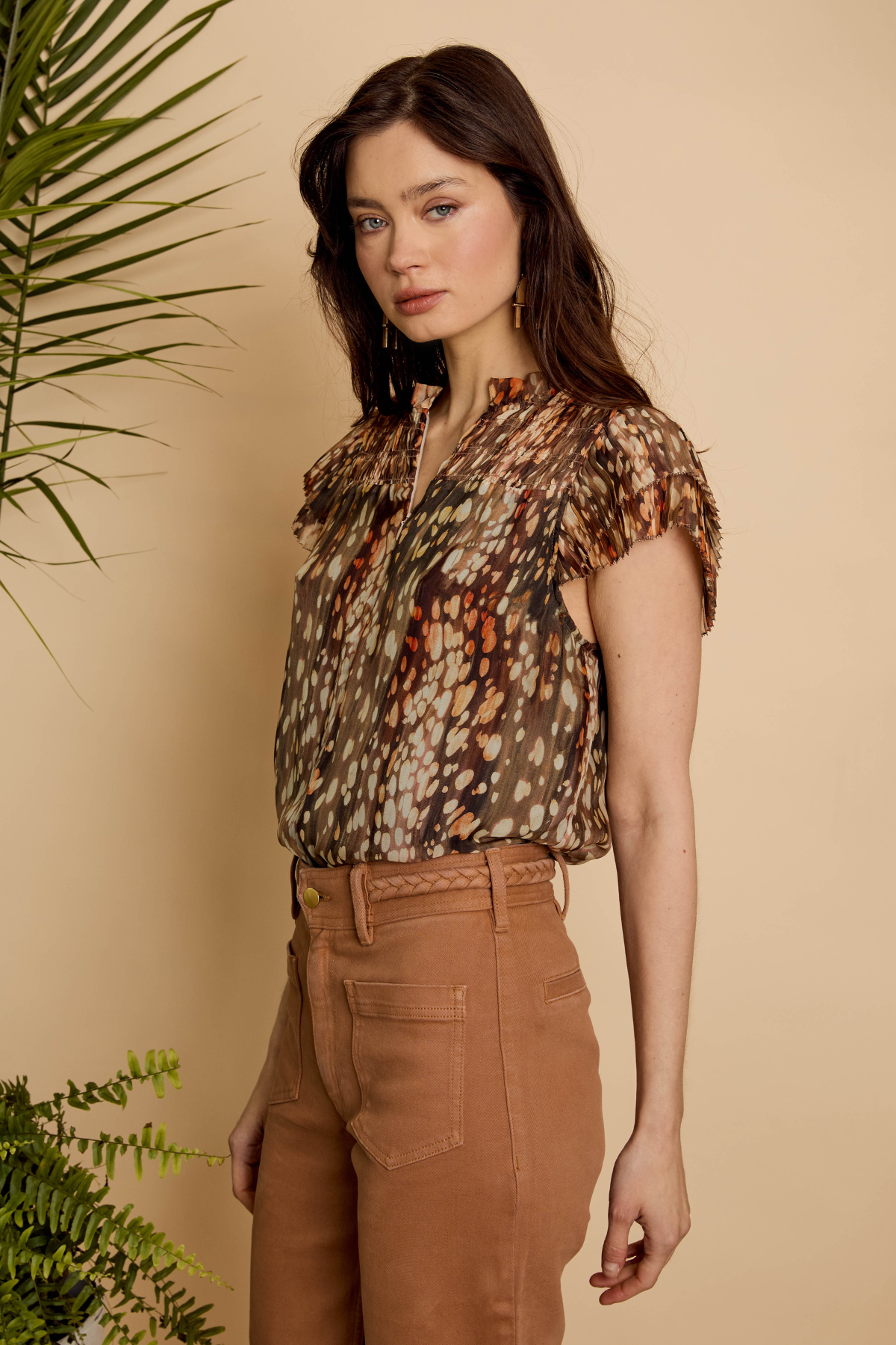 A woman modeling a short-sleeve button-up blouse with an abstract animal-like print in brown, beige, and orange tones, paired with brown pants, set against a warm beige background with green plants.