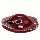 An open burgundy leather clutch bag with gold-tone hardware, showing a burgundy velvet interior. The bag is styled with white sunglasses, keys, and a white earbud case on a plain white background.