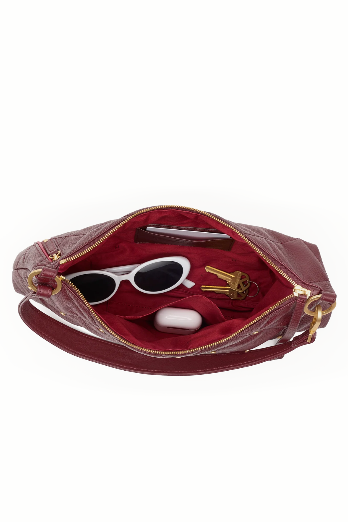 An open burgundy leather clutch bag with gold-tone hardware, showing a burgundy velvet interior. The bag is styled with white sunglasses, keys, and a white earbud case on a plain white background.