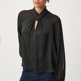 A woman wearing a black long-sleeve blouse with sheer mesh sleeves and a gathered keyhole-style neckline, paired with dark patterned pants, against a plain light gray studio background.