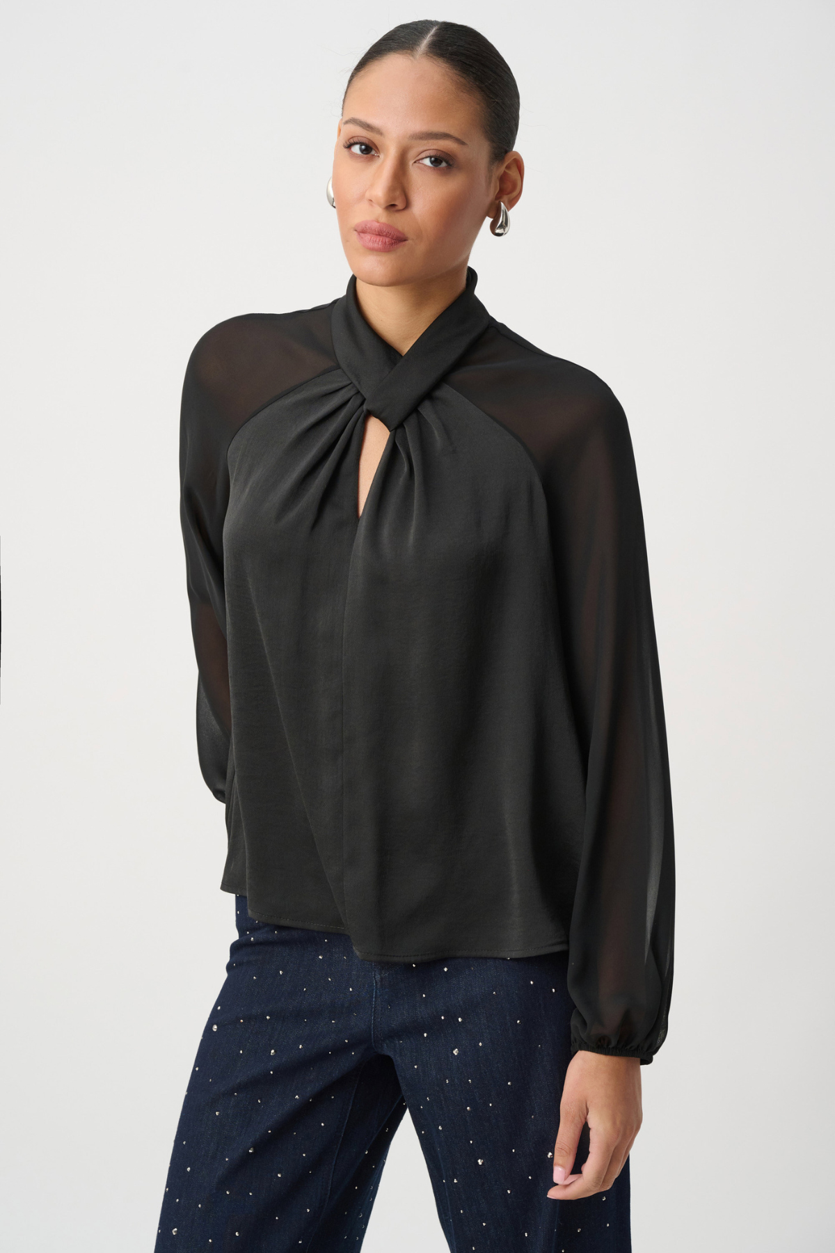 A woman wearing a black long-sleeve blouse with sheer mesh sleeves and a gathered keyhole-style neckline, paired with dark patterned pants, against a plain light gray studio background.