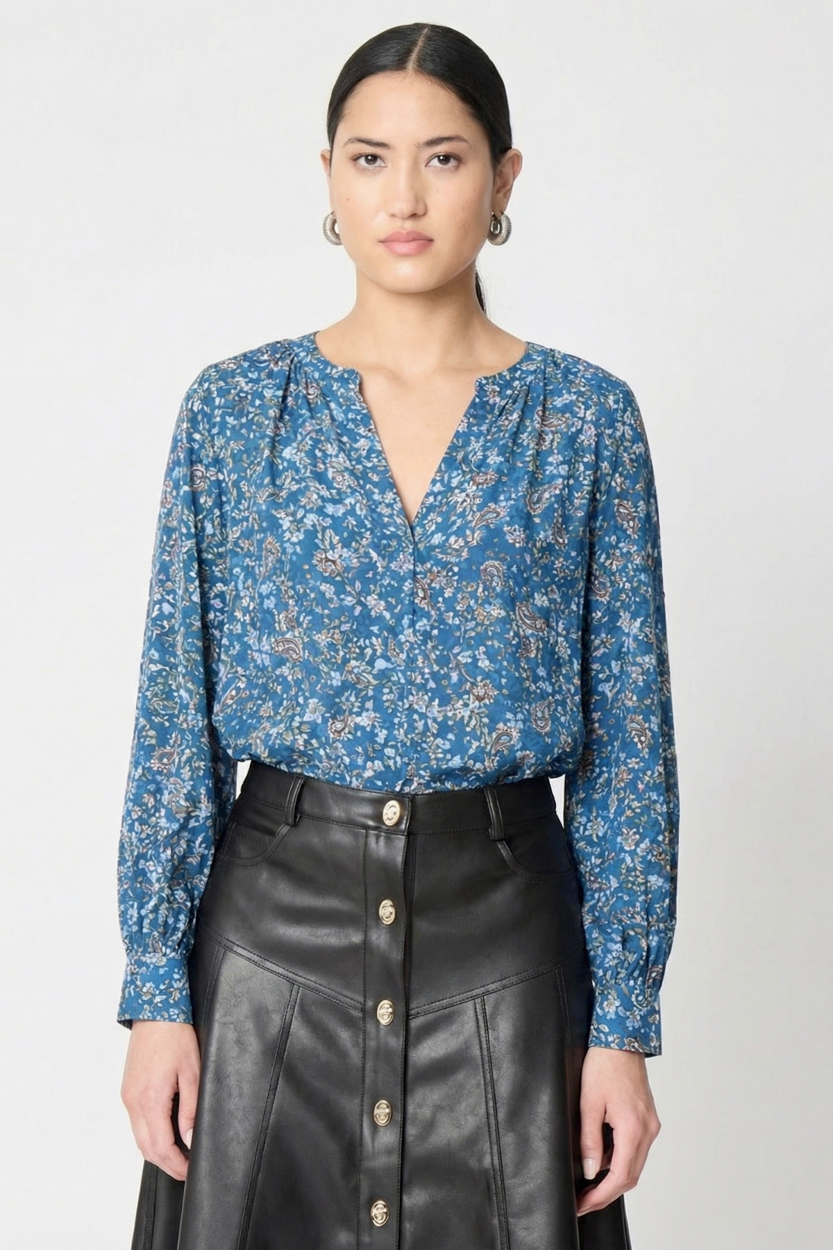 model wearing a long sleeve printed blue blouse with a v neck line and a black leather skirt with buttons against a white background