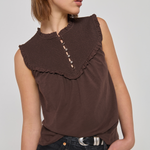 A woman wearing a sleeveless brown top with a wide ruffled collar and decorative buttons down the center, paired with dark jeans, against a plain light gray studio background.