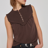 A woman wearing a sleeveless brown top with a wide ruffled collar and decorative buttons down the center, paired with dark jeans, against a plain light gray studio background.