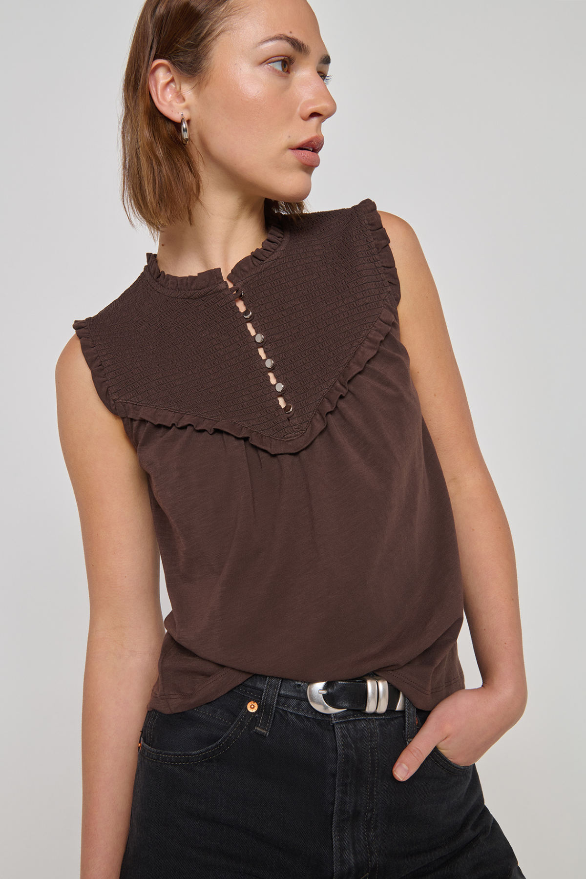 A woman wearing a sleeveless brown top with a wide ruffled collar and decorative buttons down the center, paired with dark jeans, against a plain light gray studio background.