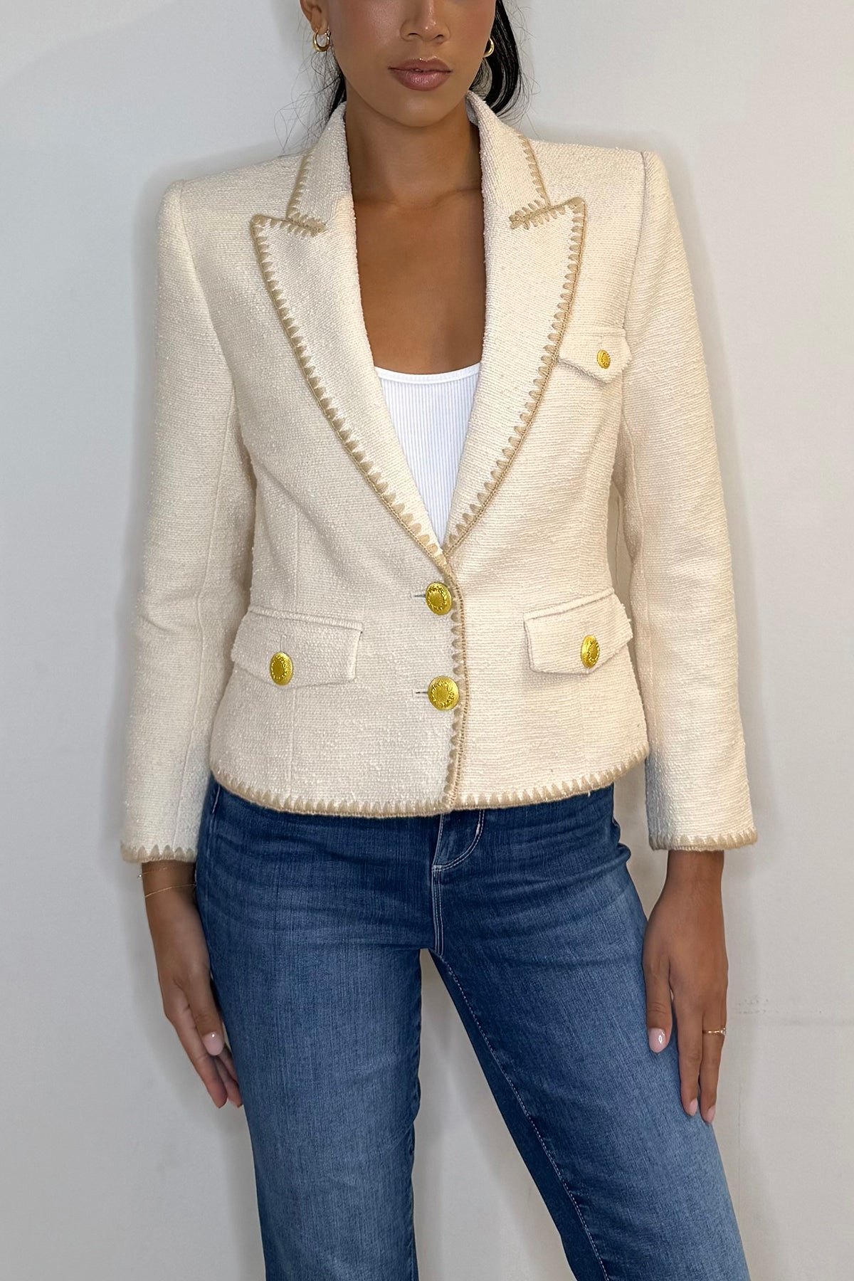Woman wearing a cream blazer with gold buttons and blue jeans against a plain background