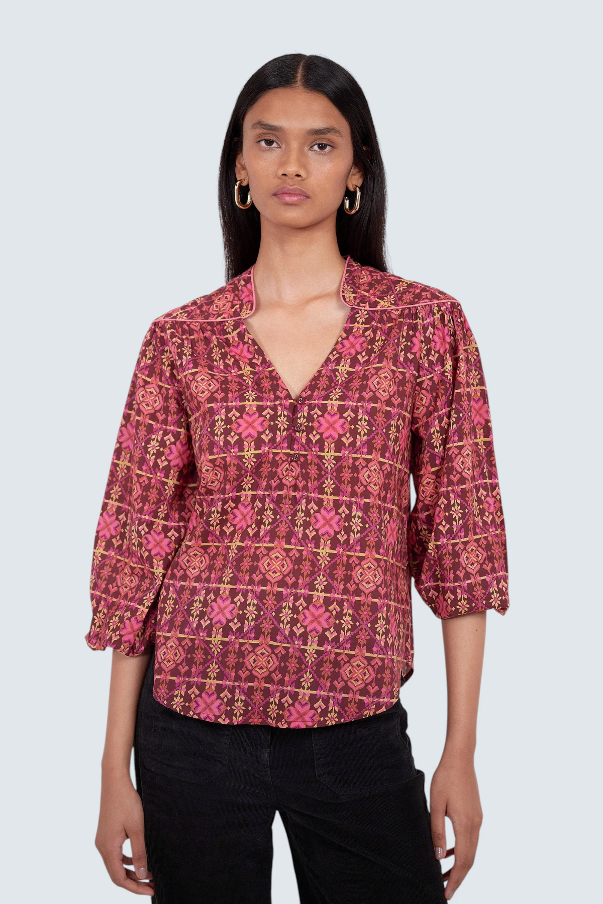 A woman modeling a patterned blouse with a split V-neck and three-quarter sleeves, paired with black pants, against a light gray studio background.