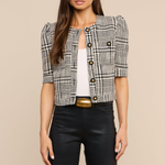 A woman modeling a short-sleeve black and white plaid tweed jacket with puff sleeves, styled over a white top and black pants with a gold belt, against a solid beige background.