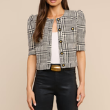 A woman modeling a short-sleeve black and white plaid tweed jacket with puff sleeves, styled over a white top and black pants with a gold belt, against a solid beige background.