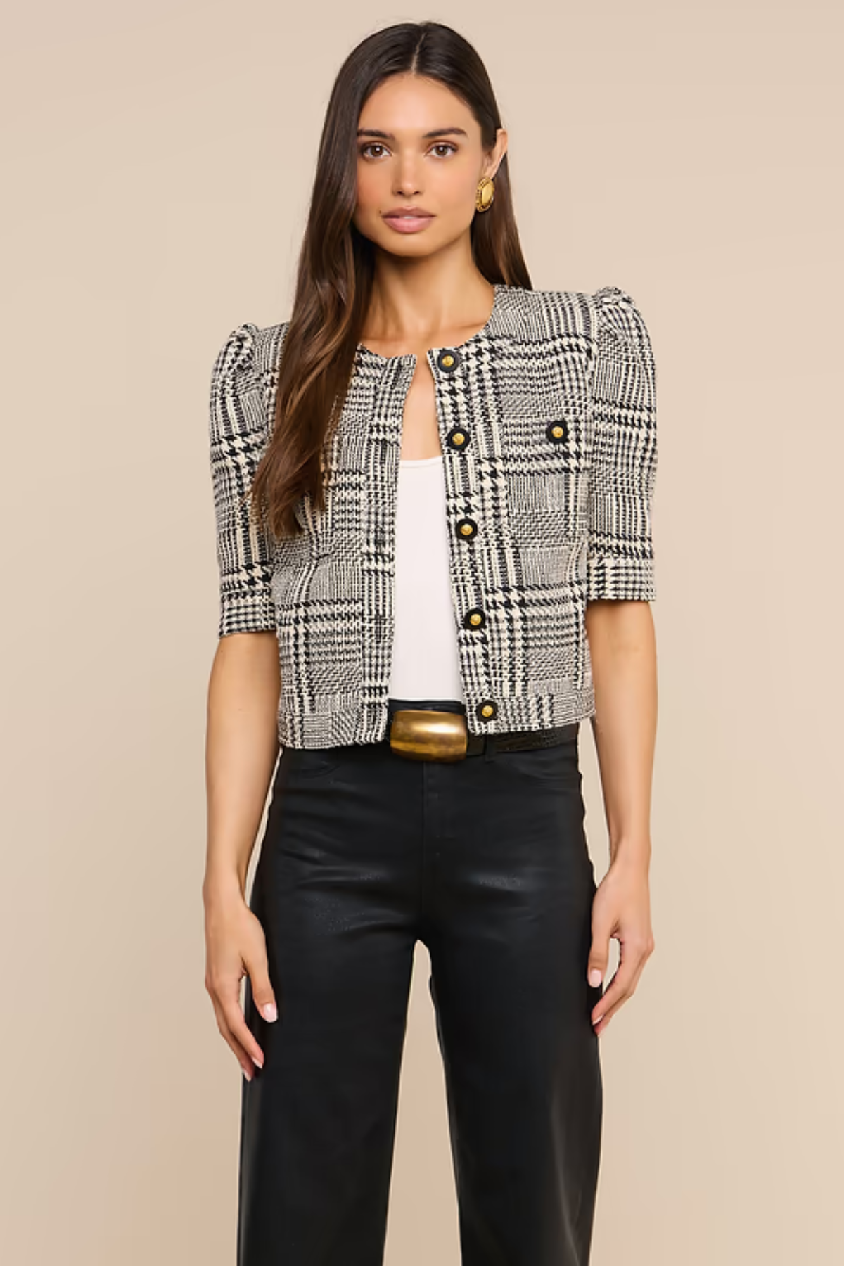 A woman modeling a short-sleeve black and white plaid tweed jacket with puff sleeves, styled over a white top and black pants with a gold belt, against a solid beige background.