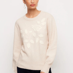 A woman modeling a cream-colored knit sweater with a raised floral and leaf pattern on the front, paired with dark navy pants, against a plain white studio background.