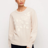 A woman modeling a cream-colored knit sweater with a raised floral and leaf pattern on the front, paired with dark navy pants, against a plain white studio background.