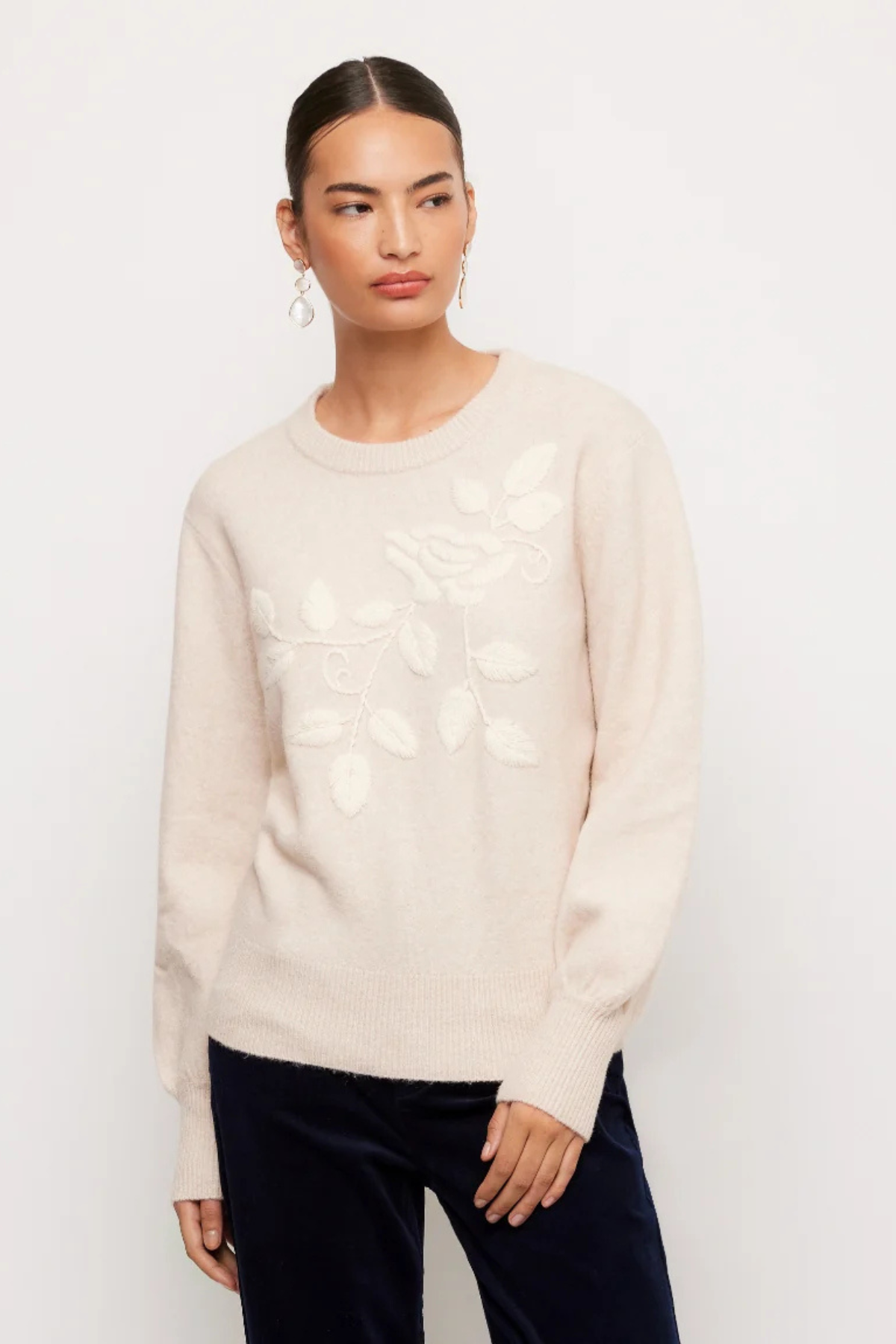 A woman modeling a cream-colored knit sweater with a raised floral and leaf pattern on the front, paired with dark navy pants, against a plain white studio background.