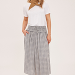 A woman modeling a white short-sleeve top with a brown neck scarf, paired with a high-waisted gray and white plaid midi skirt and tan ankle boots, against a plain light beige studio background.