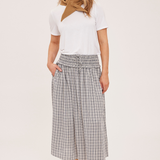 A woman modeling a white short-sleeve top with a brown neck scarf, paired with a high-waisted gray and white plaid midi skirt and tan ankle boots, against a plain light beige studio background.