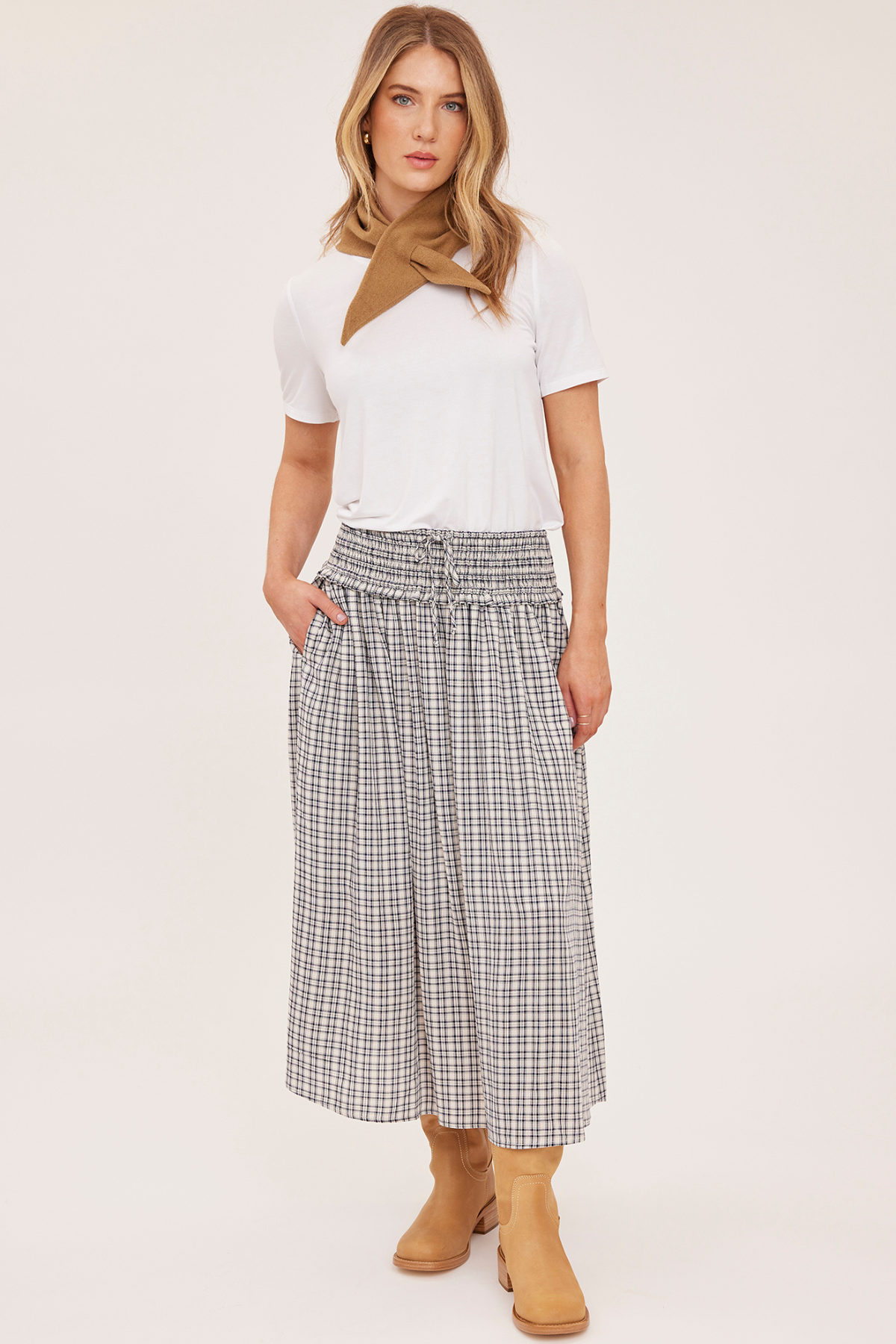 A woman modeling a white short-sleeve top with a brown neck scarf, paired with a high-waisted gray and white plaid midi skirt and tan ankle boots, against a plain light beige studio background.