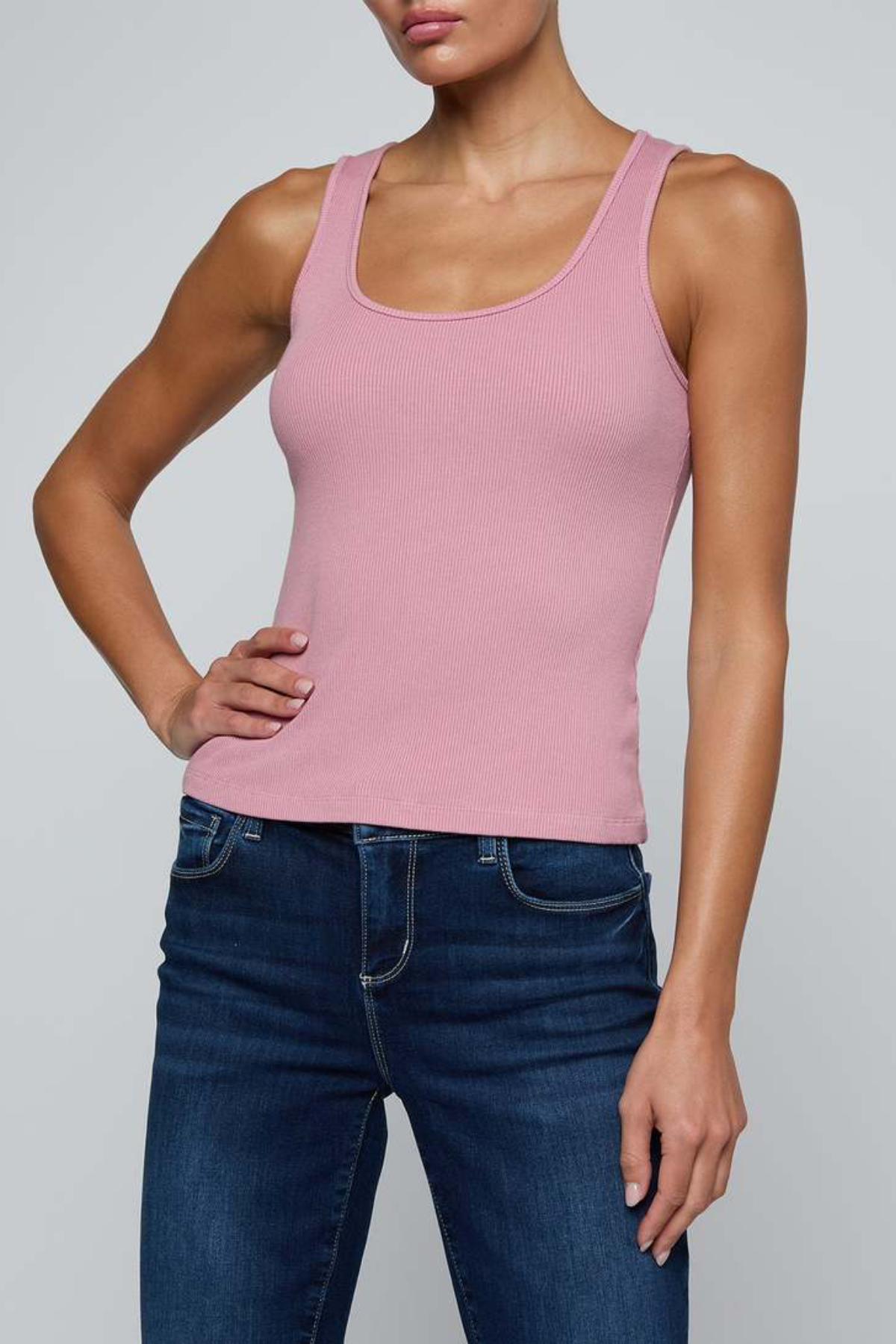 A woman wearing a fitted scoop-neck tank top in dusty pink with dark blue jeans, shown from the nose down against a light gray studio background.