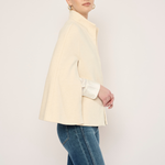 A woman modeling a cream-colored structured jacket with wide sleeves, paired with blue jeans, against a plain off-white studio background.