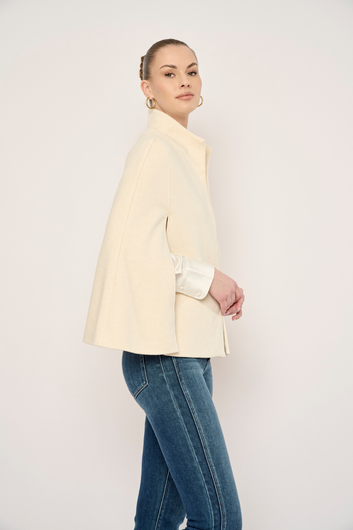 A woman modeling a cream-colored structured jacket with wide sleeves, paired with blue jeans, against a plain off-white studio background.