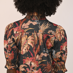 Back view of a person wearing a short-sleeve blouse with a dark tropical floral print in red, beige, and brown tones, featuring decorative beaded trim at the collar and cuffs.