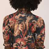 Back view of a person wearing a short-sleeve blouse with a dark tropical floral print in red, beige, and brown tones, featuring decorative beaded trim at the collar and cuffs.