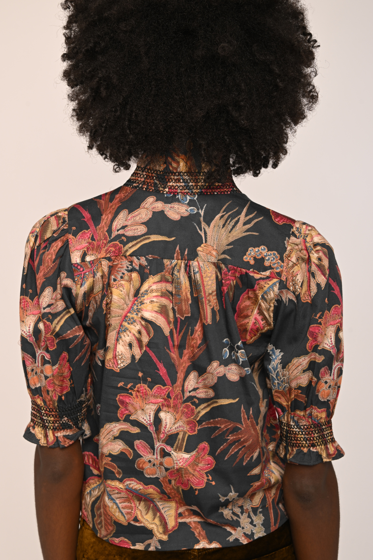 Back view of a person wearing a short-sleeve blouse with a dark tropical floral print in red, beige, and brown tones, featuring decorative beaded trim at the collar and cuffs.