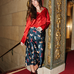 Woman in a red blouse and patterned skirt standing in front of an ornate wall.