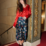 Woman in a red blouse and patterned skirt standing in front of an ornate wall.