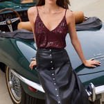 A woman posing in front of a classic green convertible car, wearing a burgundy sequined spaghetti-strap top and a black faux-leather midi skirt with a silver button-down front.
