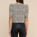 A woman shown from the back wearing a black and white plaid cropped jacket with puff sleeves and black wide-leg trousers, set against a solid beige background.