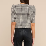 A woman shown from the back wearing a black and white plaid cropped jacket with puff sleeves and black wide-leg trousers, set against a solid beige background.