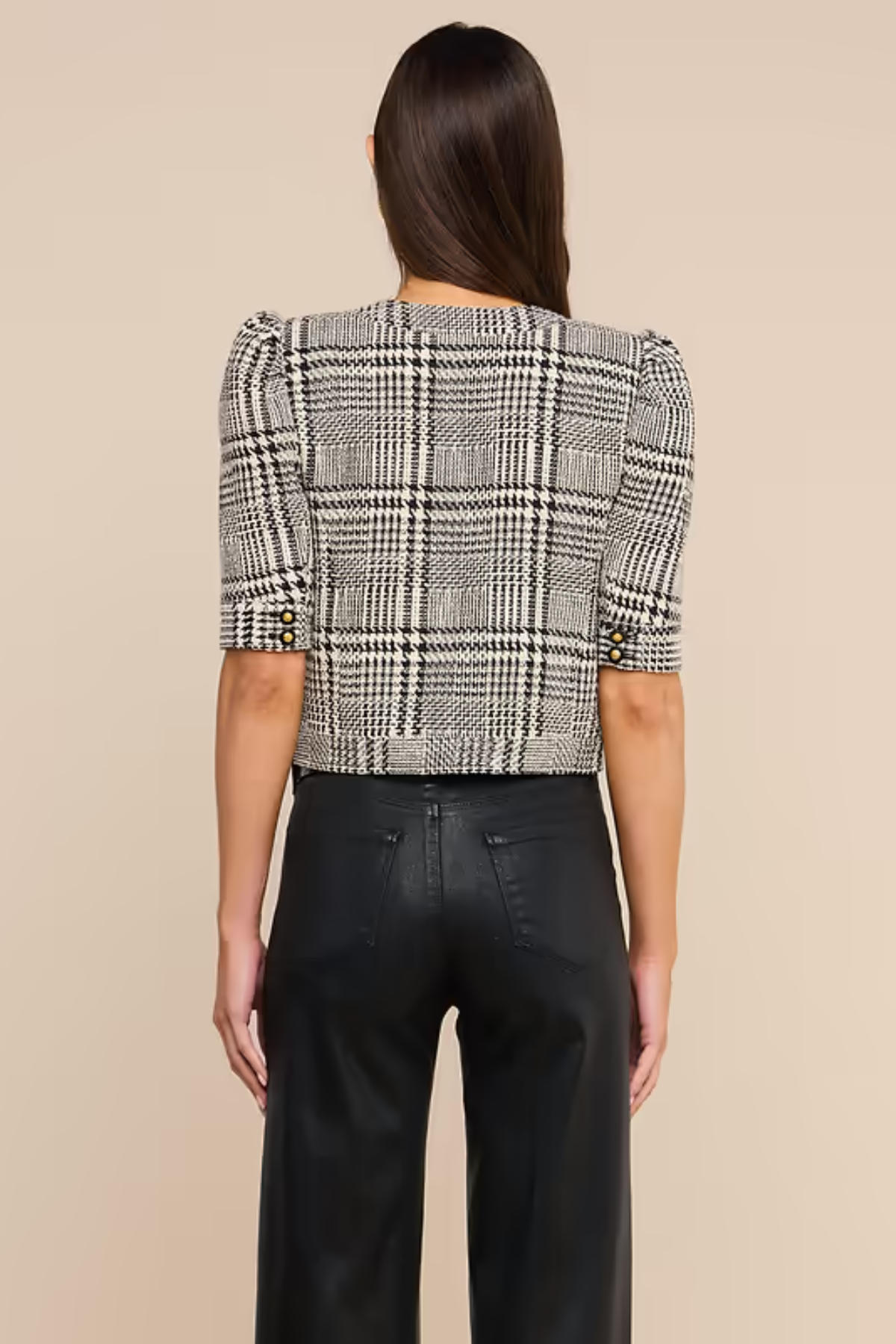 A woman shown from the back wearing a black and white plaid cropped jacket with puff sleeves and black wide-leg trousers, set against a solid beige background.