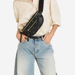 A model wearing a cream-colored shirt and light-wash blue jeans, carrying a black faux-leather fanny pack with gold studs and a wide strap across their waist.