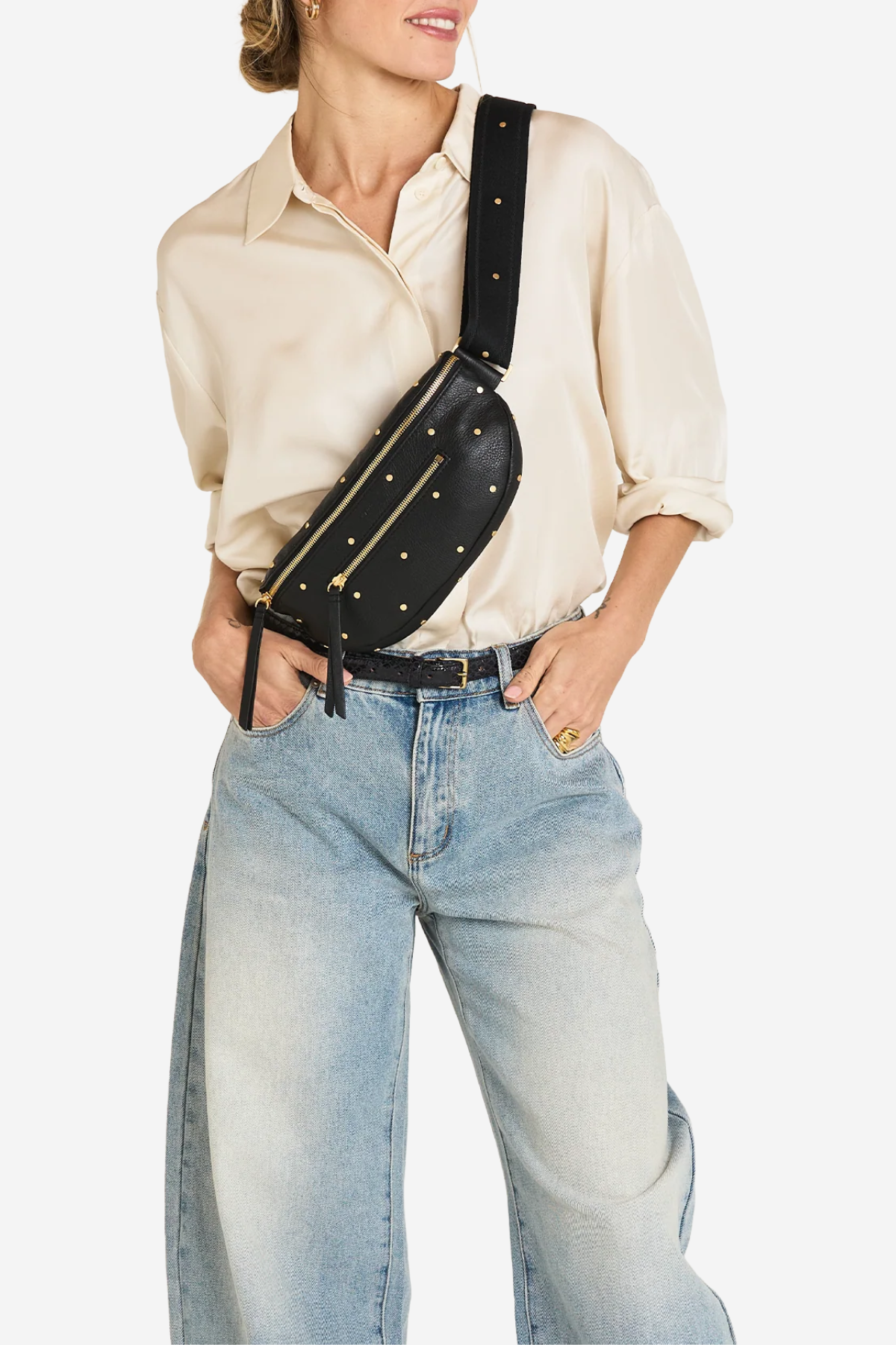 A model wearing a cream-colored shirt and light-wash blue jeans, carrying a black faux-leather fanny pack with gold studs and a wide strap across their waist.