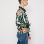 A woman in profile wearing a green long-sleeve button-up shirt with a detailed beige and brown paisley pattern, paired with blue jeans, against a plain white studio background.