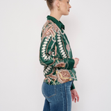 A woman in profile wearing a green long-sleeve button-up shirt with a detailed beige and brown paisley pattern, paired with blue jeans, against a plain white studio background.