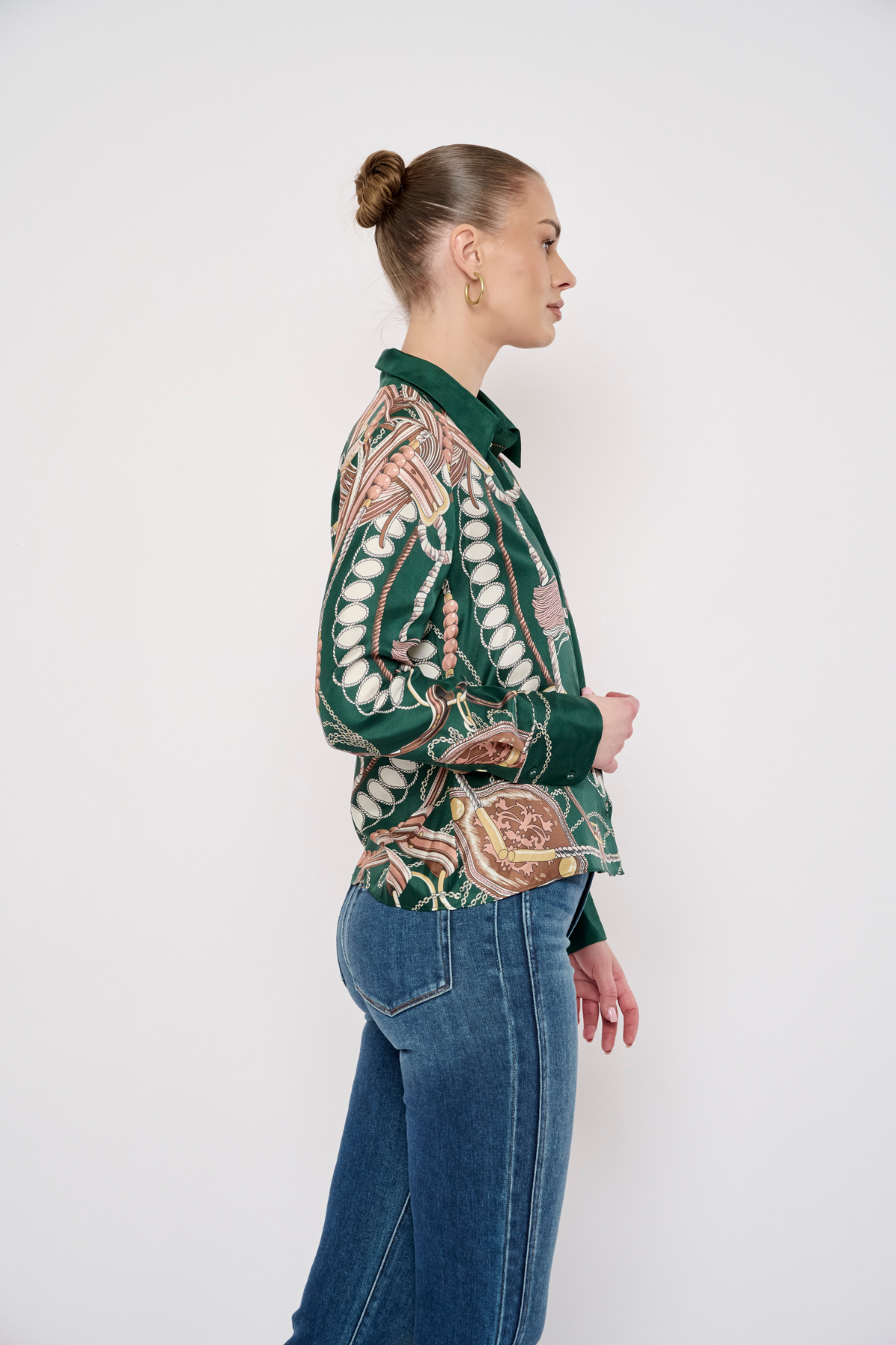 A woman in profile wearing a green long-sleeve button-up shirt with a detailed beige and brown paisley pattern, paired with blue jeans, against a plain white studio background.
