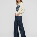 A woman modeling a cream knit sweater with a dark blue floral pattern on the chest and dark blue patterned wide-leg trousers, standing against a light gray studio background.