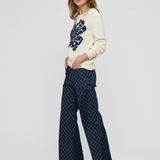 A woman modeling a cream knit sweater with a dark blue floral pattern on the chest and dark blue patterned wide-leg trousers, standing against a light gray studio background.