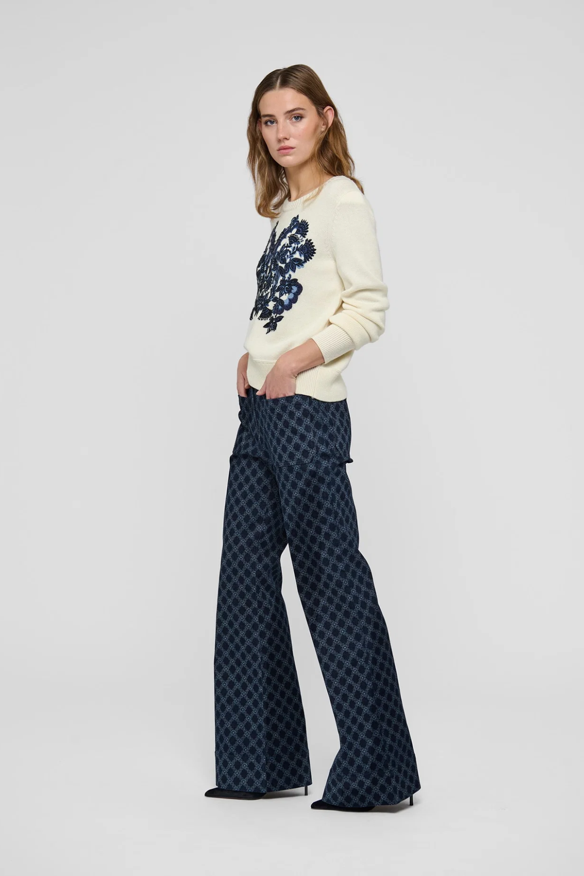 A woman modeling a cream knit sweater with a dark blue floral pattern on the chest and dark blue patterned wide-leg trousers, standing against a light gray studio background.