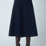 A dark navy blue A-line midi skirt shown on a model from the waist down, paired with brown leather knee-high boots against a light gray studio background.