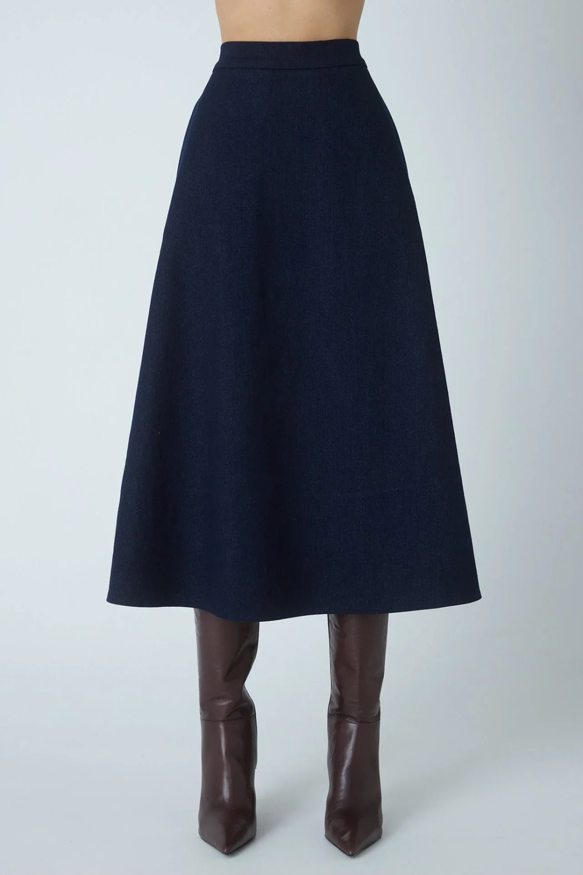 A dark navy blue A-line midi skirt shown on a model from the waist down, paired with brown leather knee-high boots against a light gray studio background.