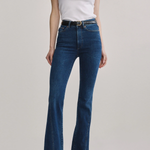 A model wearing a pair of dark blue high-waisted flare jeans with a black belt, paired with a white sleeveless top and black shoes, against a light gray studio background.