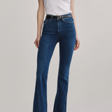 A model wearing a pair of dark blue high-waisted flare jeans with a black belt, paired with a white sleeveless top and black shoes, against a light gray studio background.