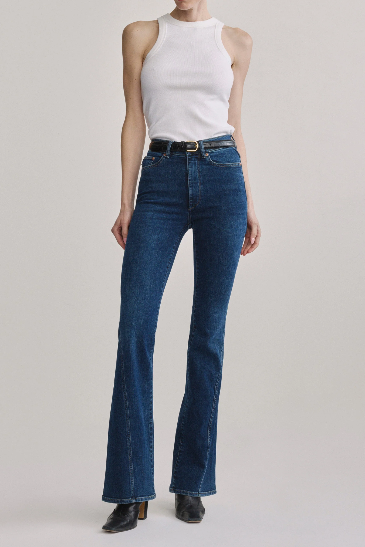 A model wearing a pair of dark blue high-waisted flare jeans with a black belt, paired with a white sleeveless top and black shoes, against a light gray studio background.