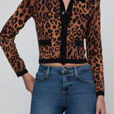 A woman wearing a cropped leopard-print cardigan with a black collar and front buttons, paired with blue jeans, against a light gray studio background.
